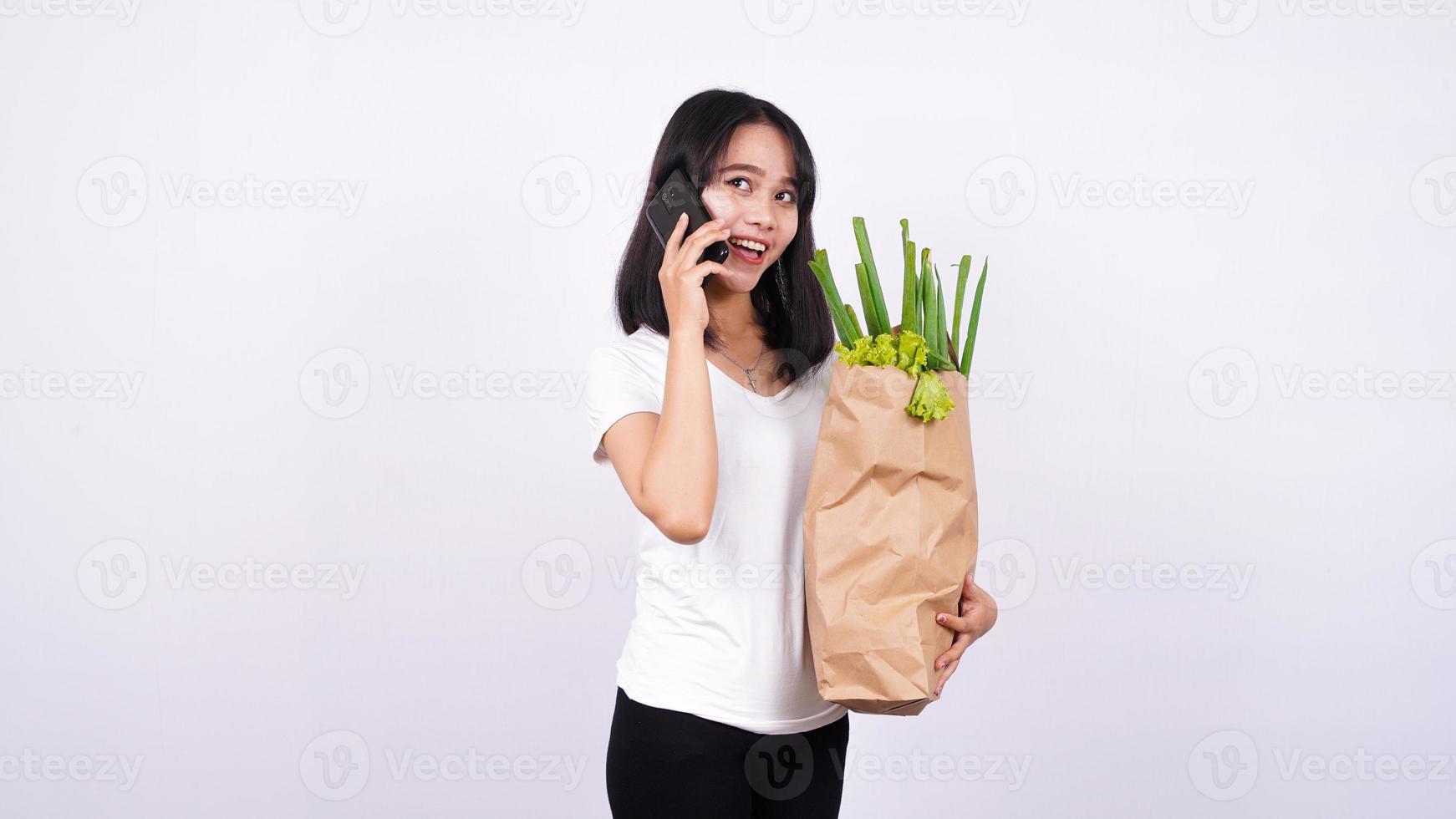 Aziatische vrouw met papieren zak verse groenten en praten aan een telefoon met geïsoleerde witte achtergrond foto