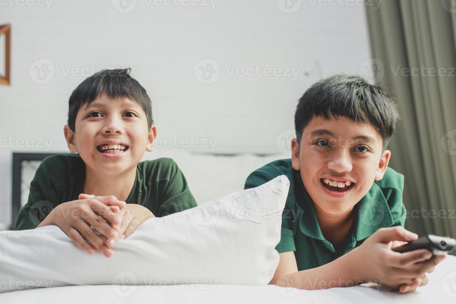 twee broers die op het bed liggen en glimlachen tijdens het tv-kijken en een afstandsbediening vasthouden foto