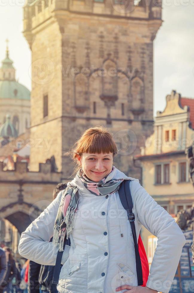 jonge meisjestoerist met witte jas kijkt naar de camera en lacht naar charles bridge karluv most in praag historisch centrum foto