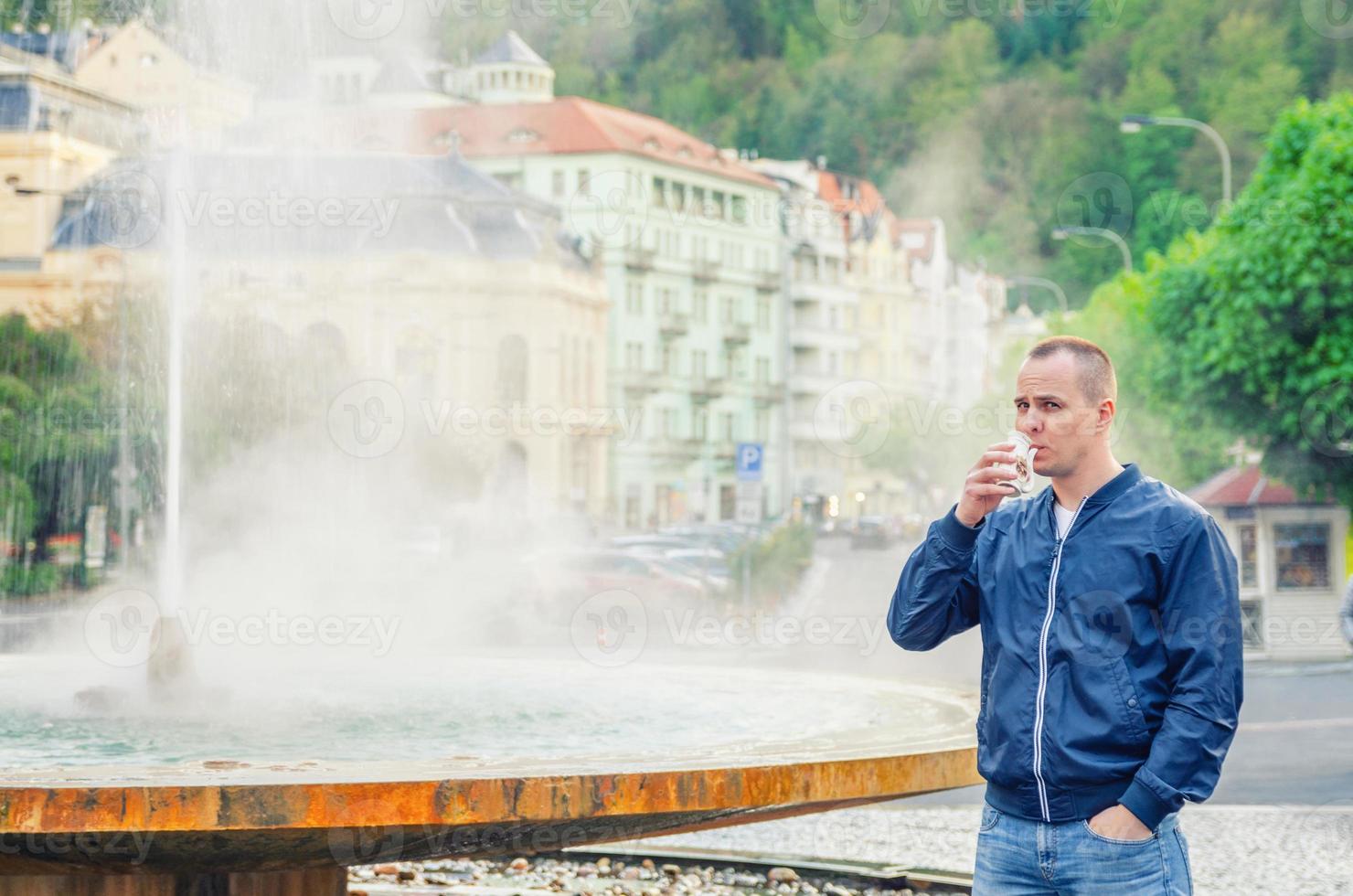 jonge man toerist met blauwe jas kijkt naar de camera en drinkt therapeutisch mineraalwater uit de beker foto