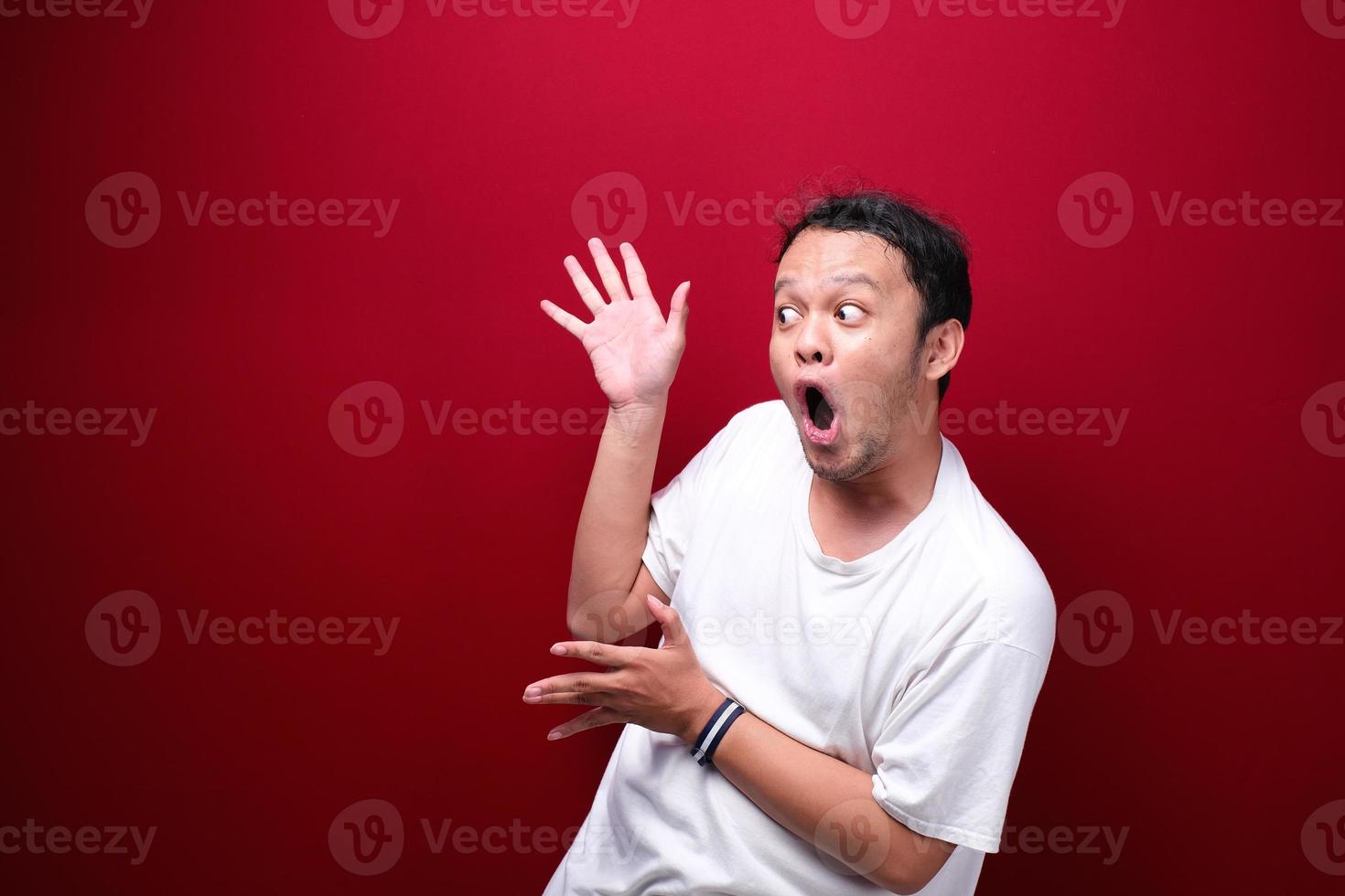 jonge aziatische man is verrast en schreeuwt wow met naar rechts te wijzen met zijn hand geïsoleerd op rode achtergrond. foto