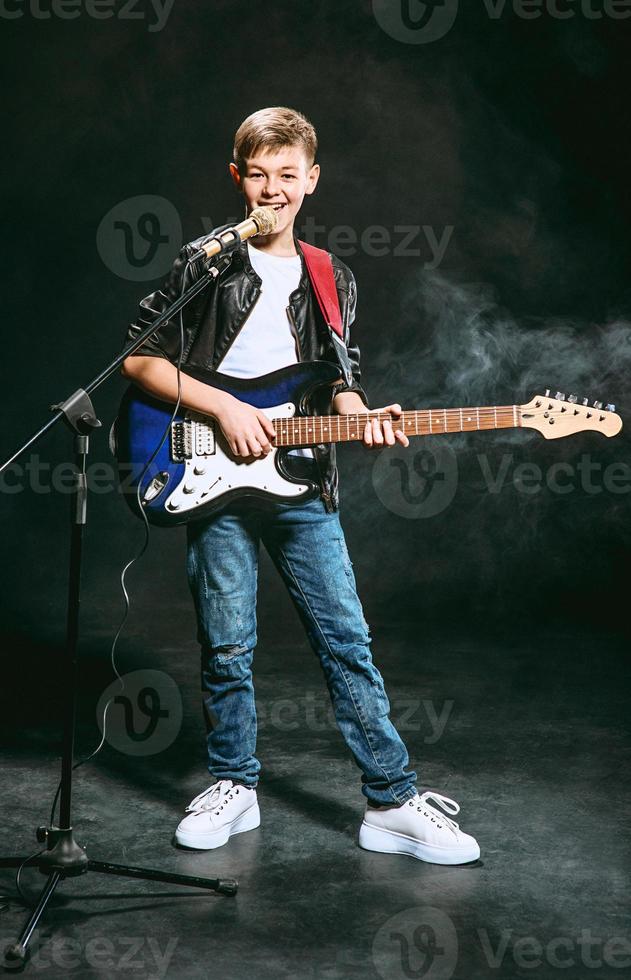 portret van blanke tiener in wit t-shirt, spijkerbroek en leren jas met microfoon en gitaar zingen op donkere achtergrond. hobby en glorie concept foto