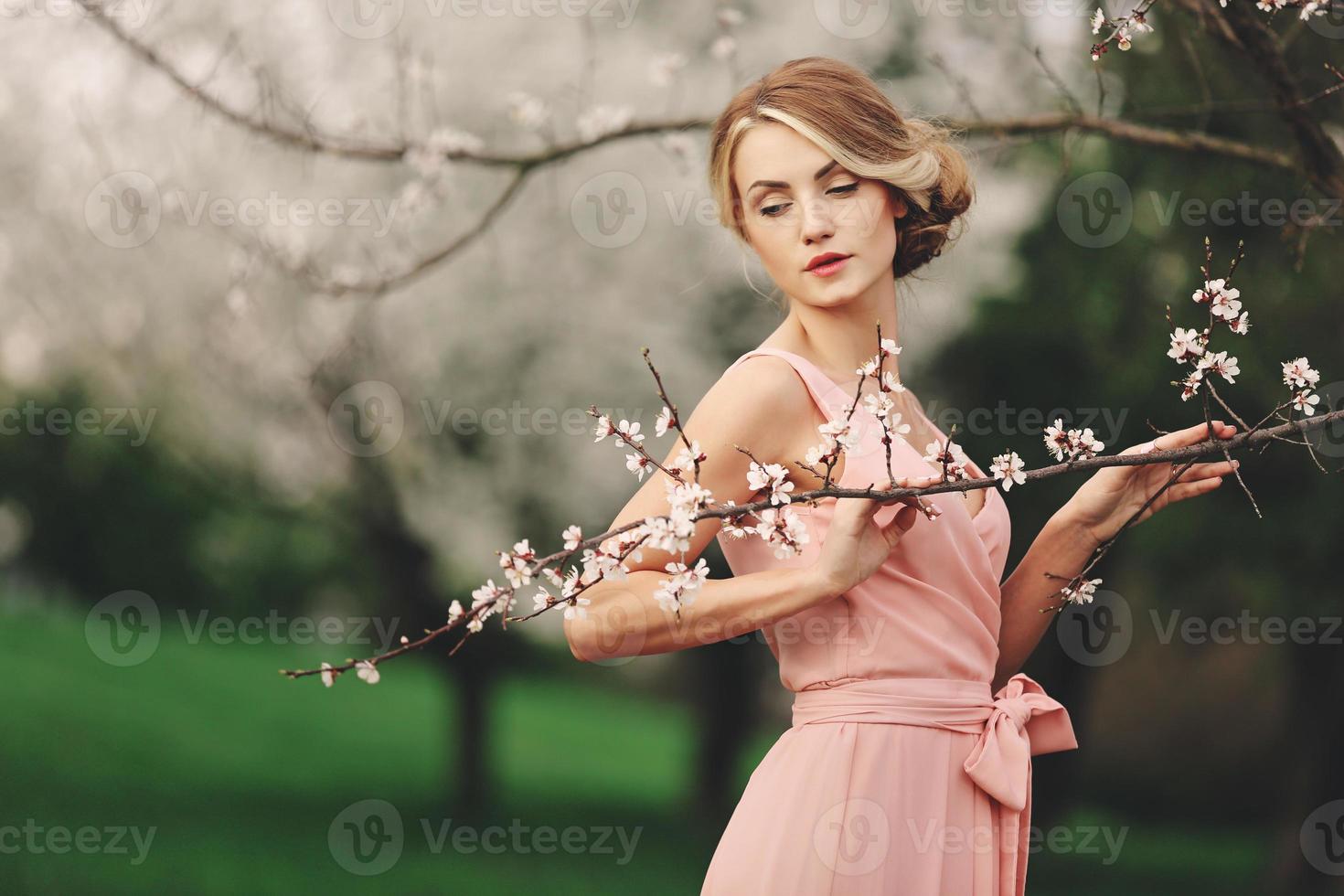 jonge stijlvolle vrouw in de buurt van bloeiende bloeiende boom in het park. lente achtergrond. blond meisje met kapsel in roze jurk. ruimte kopiëren. foto