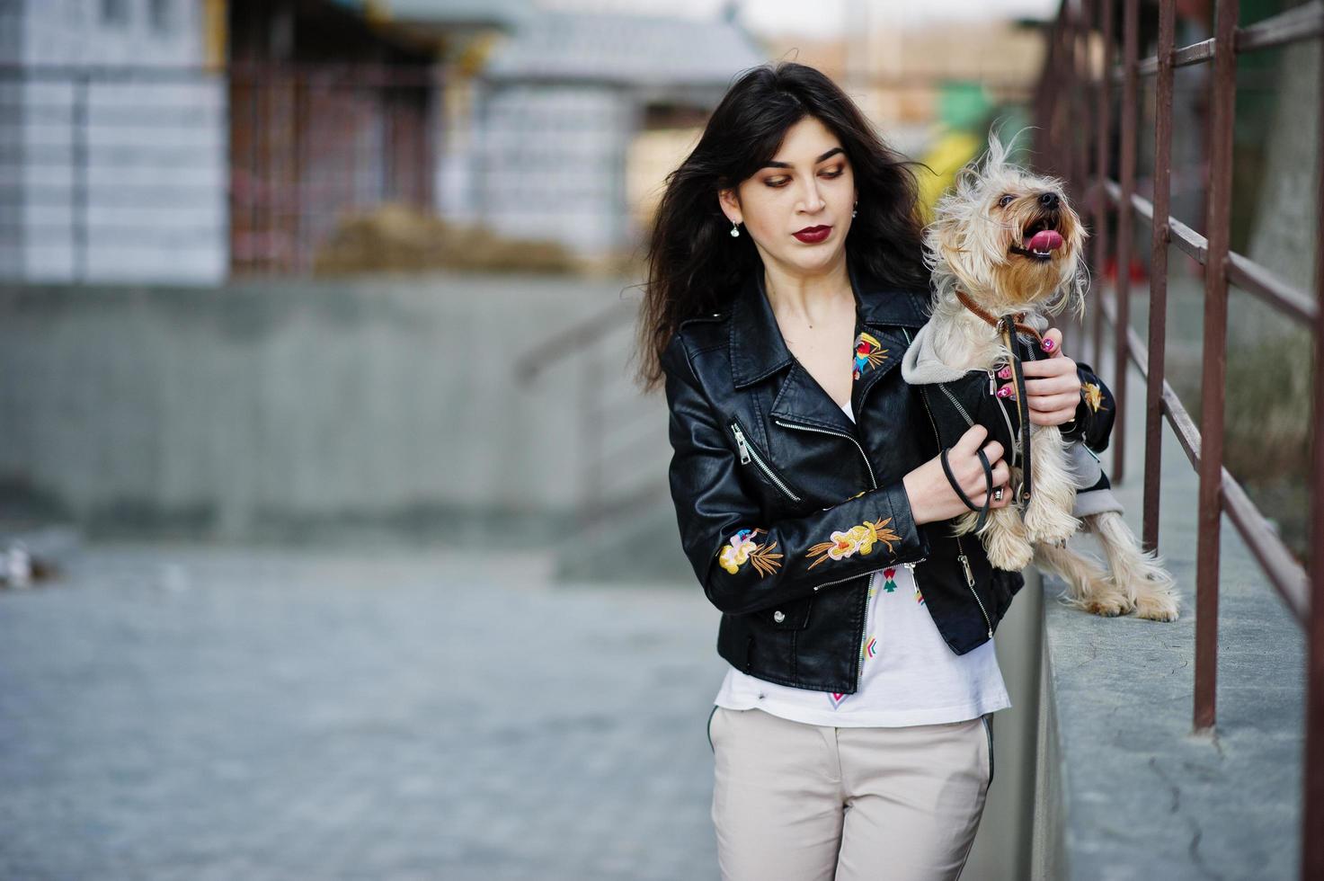 brunette zigeunermeisje met yorkshire terrier hond gesteld tegen stalen balustrades. model slijtage op leren jas en t-shirt met ornament, broek. foto