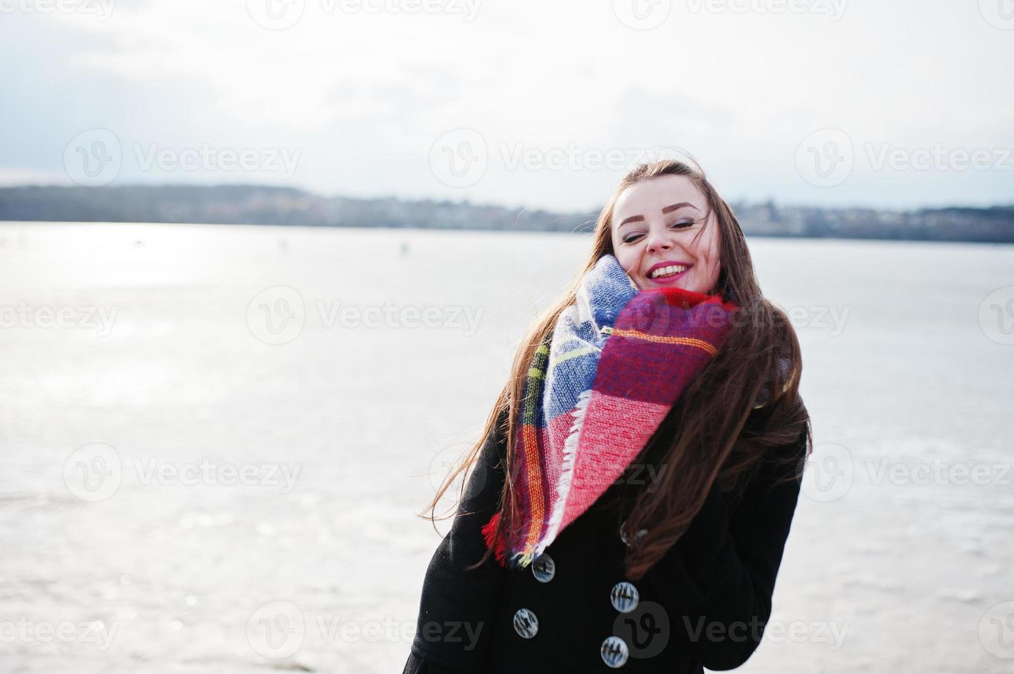 casual jong meisje op zwarte jas, sjaal en muts tegen bevroren rivier op zonnig winterweer. foto
