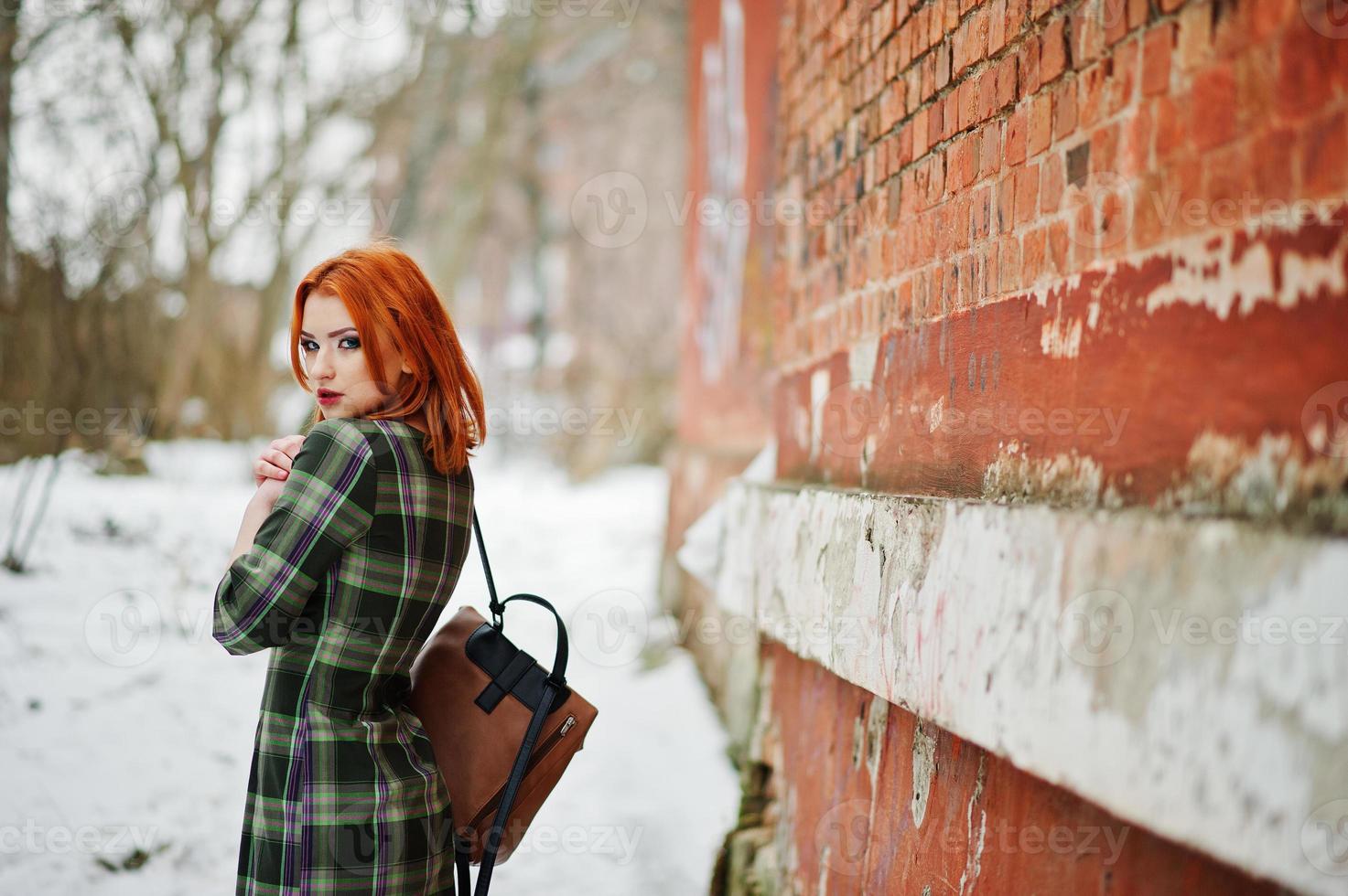 een openluchtportret van een jong mooi meisje met rood haar die geruite kleding met girly rugzakken dragen die zich op de bakstenen muurachtergrond in de winterdag bevinden. foto