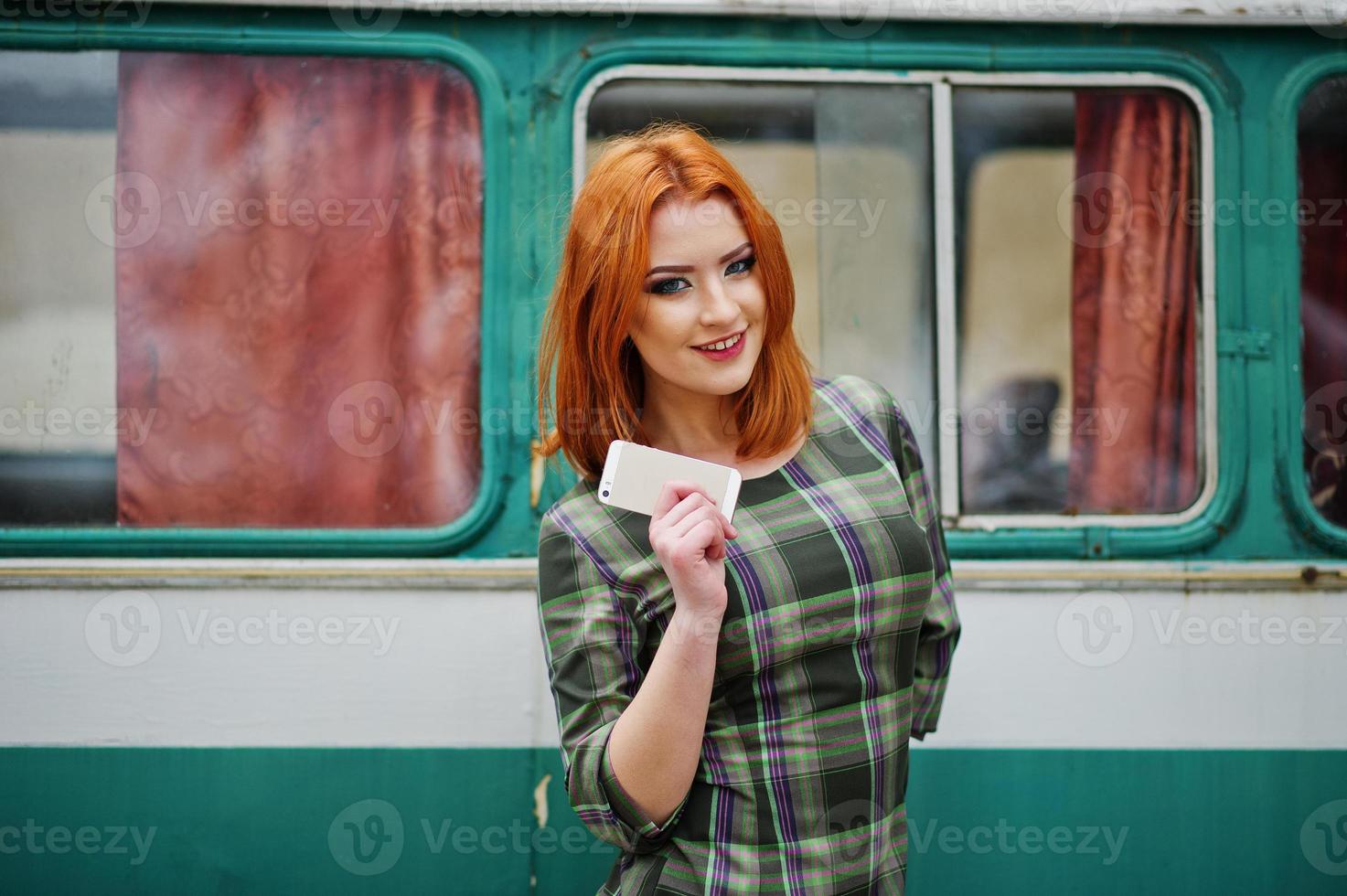 jonge roodharige meisje dame met mobiele telefoon en koptelefoon, gekleed op geruite jurk achtergrond oude vintage turqoise minivan bus. foto