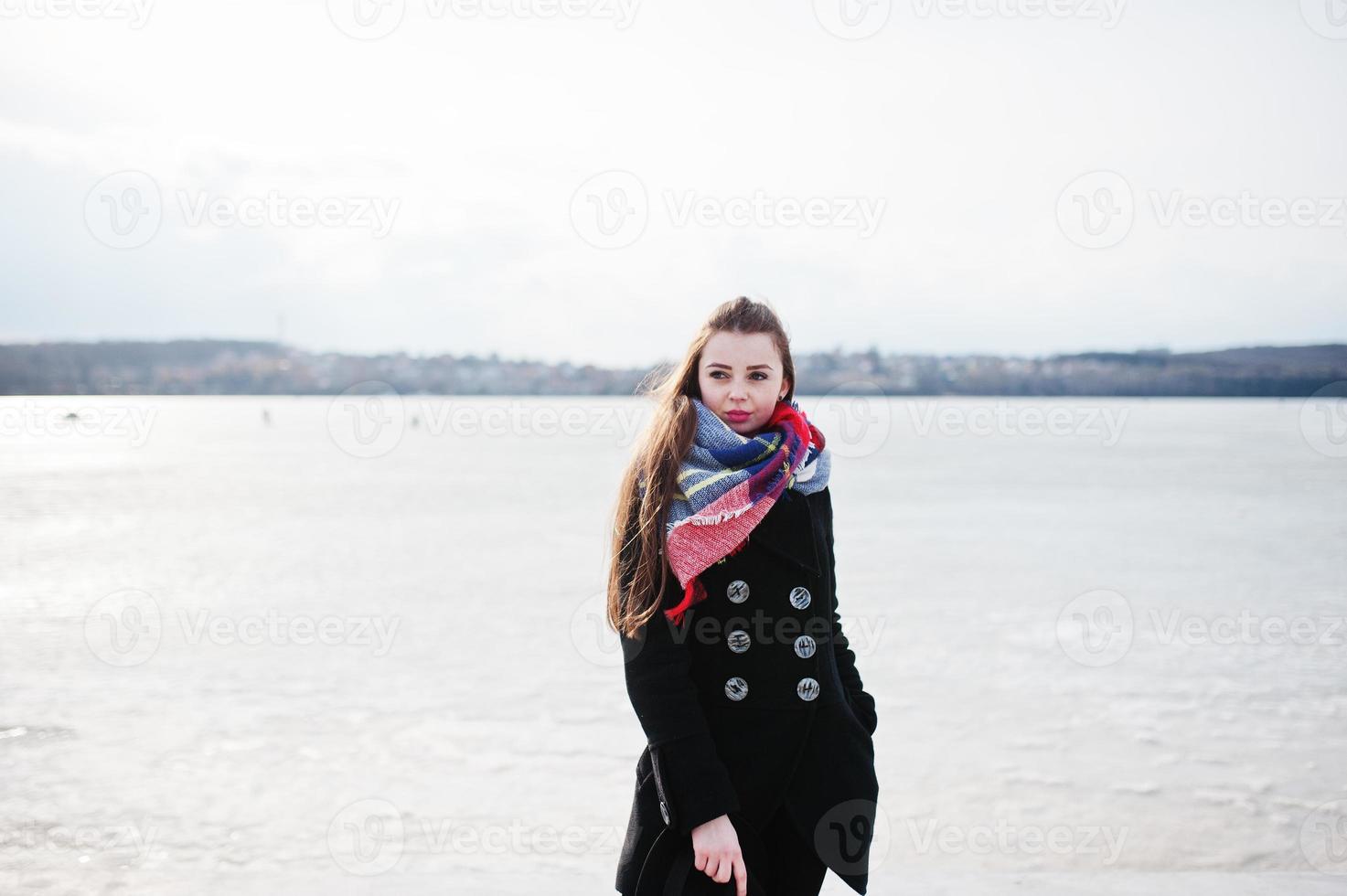 casual jong meisje op zwarte jas, sjaal en muts tegen bevroren rivier op zonnig winterweer. foto