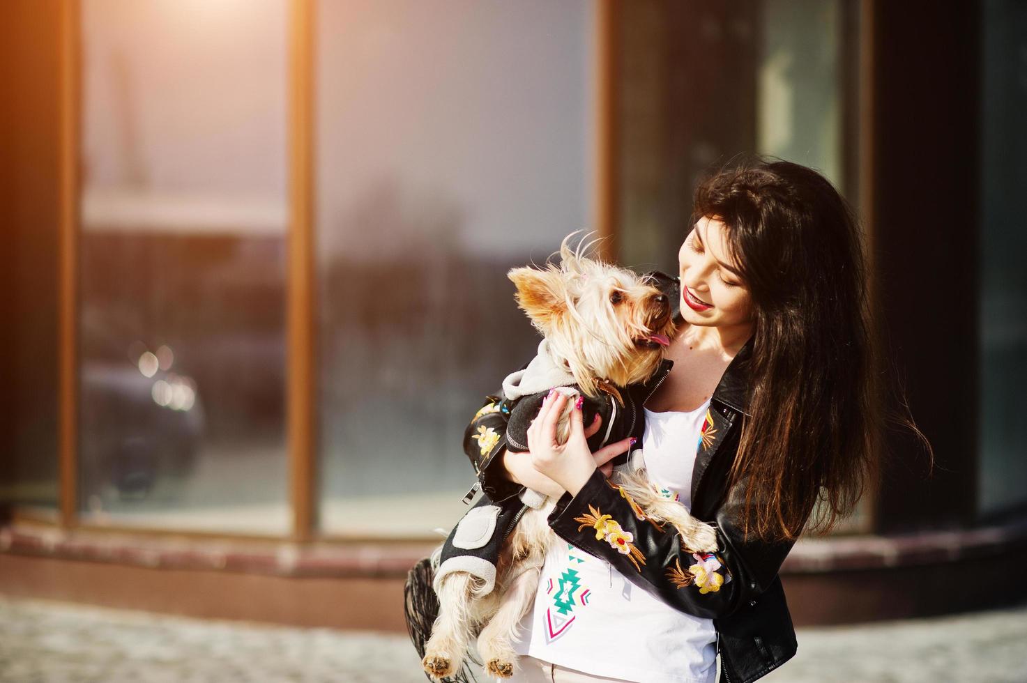 brunette zigeunermeisje met yorkshire terrier hond gesteld tegen grote ramen huis. model slijtage op leren jas en t-shirt met ornament, broek. foto