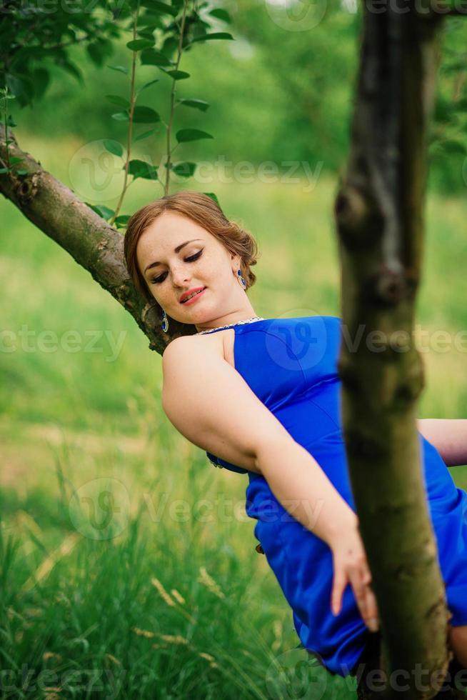 jong meisje met overgewicht op blauwe jurk poseerde achtergrond lentetuin zittend op boom. foto