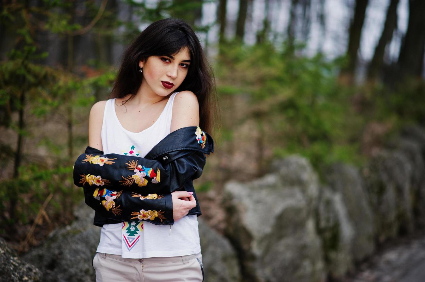 brunette zigeunermeisje gesteld tegen stenen op park. model slijtage op leren jas en t-shirt met ornament, broek. foto