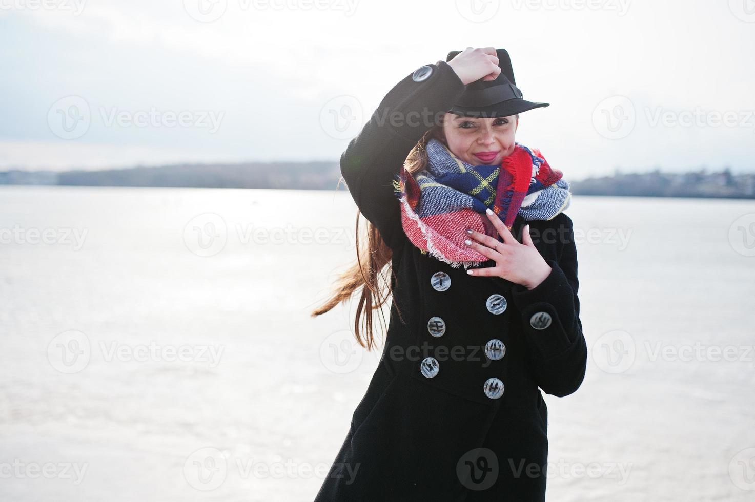 grappig casual jong meisje bij zwarte jas, sjaal en muts tegen bevroren rivier op zonnig winterweer. foto