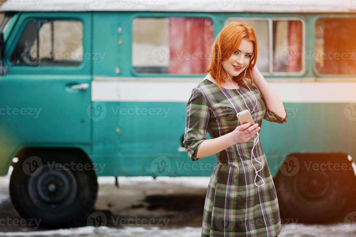 jonge roodharige meisje dame met mobiele telefoon en koptelefoon, gekleed op geruite jurk achtergrond oude vintage turqoise minivan bus. foto
