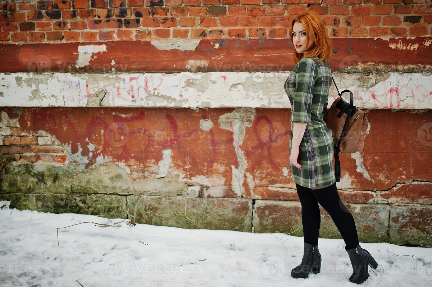 een openluchtportret van een jong mooi meisje met rood haar die geruite kleding met girly rugzakken dragen die zich op de bakstenen muurachtergrond in de winterdag bevinden. foto