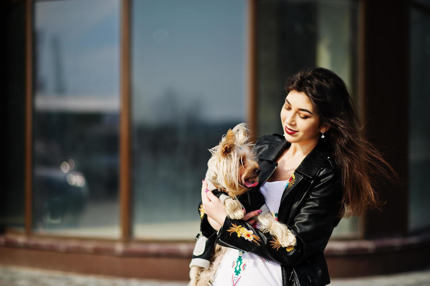 brunette zigeunermeisje met yorkshire terrier hond gesteld tegen grote ramen huis. model slijtage op leren jas en t-shirt met ornament, broek. foto