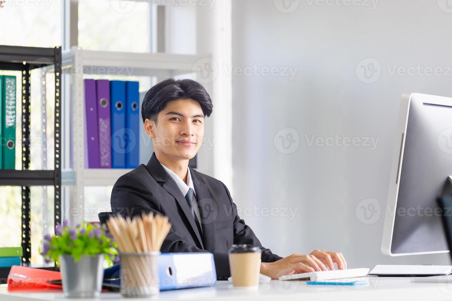 portret van een glimlachende jonge man die aan zijn bureau op kantoor zit, zakenman in pak die met een laptop op kantoor werkt foto