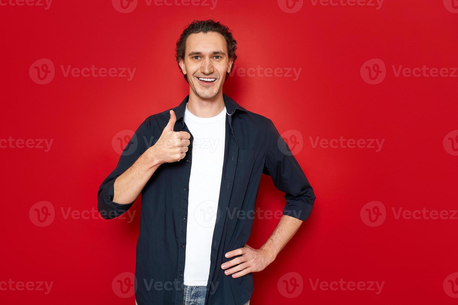 vrolijke gelukkig lachende man in blauw shirt, wit t-shirt houdt hand voor hem en toont zijn duim. geïsoleerd op rode studio achtergrond met ruimte voor tekst. concept - goedkeuring, aanbeveling, mensen foto