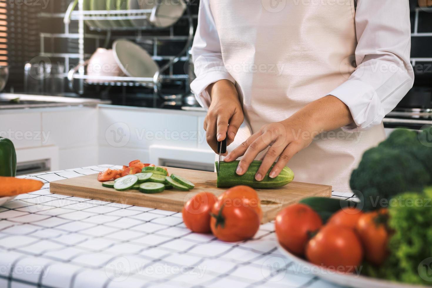 huisvrouw met mes en handen snijden komkommer op houten bord in keuken kamer. foto