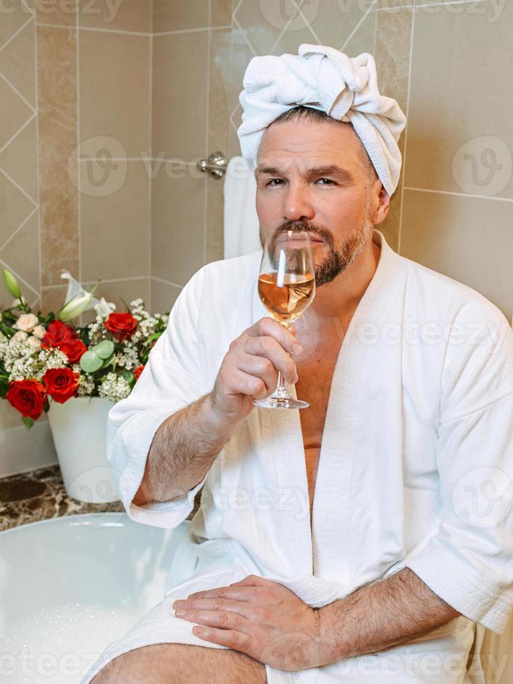 knappe volwassen man in witte badjas en handdoek op zijn hoofd zittend op de badkamer met schuim en rose wijn drinken. spa, ontspannen, levensstijl, genieten van het levensconcept. foto