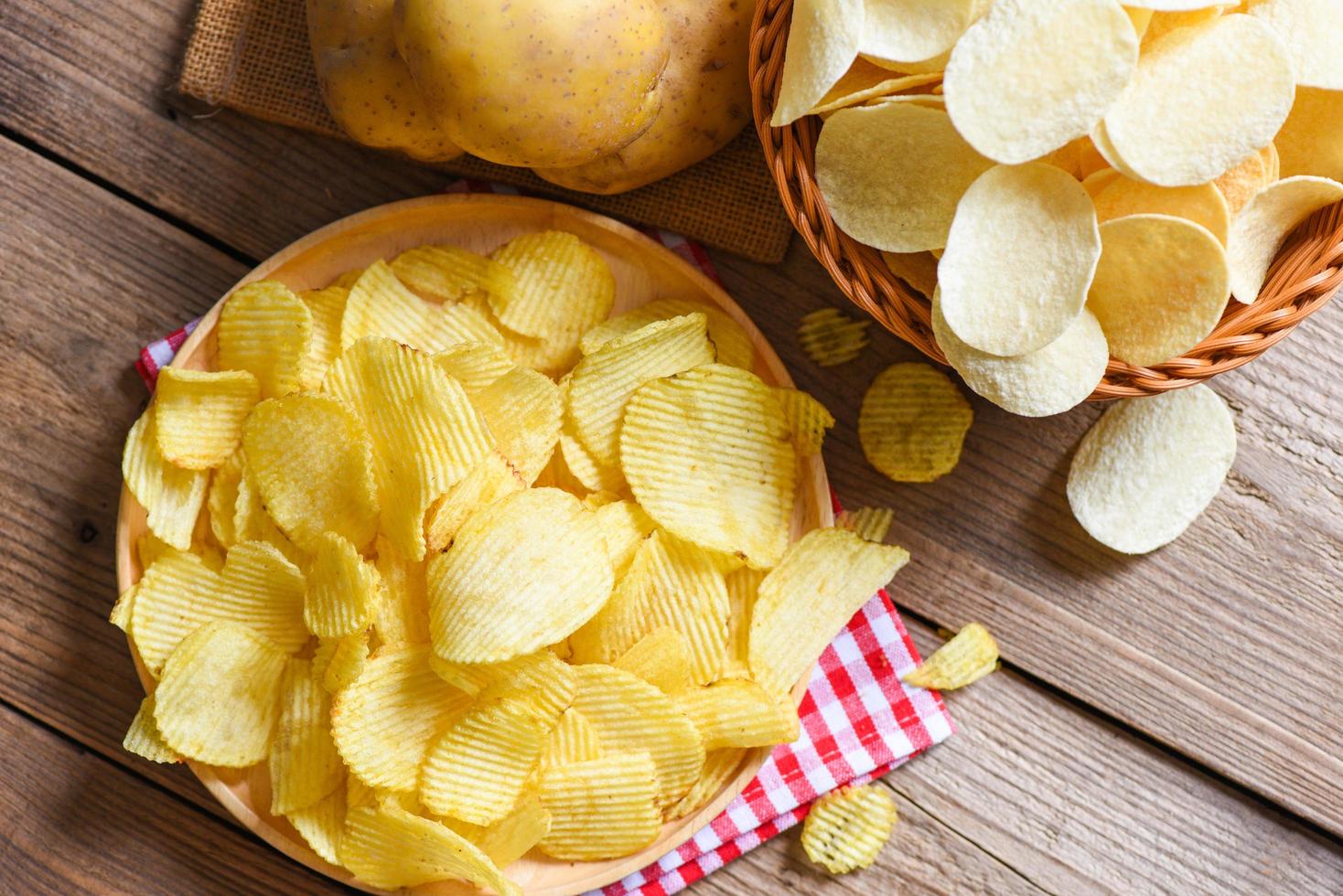 chips snack op bord, knapperige chips op de keukentafel en verse rauwe aardappelen op houten ondergrond - bovenaanzicht foto