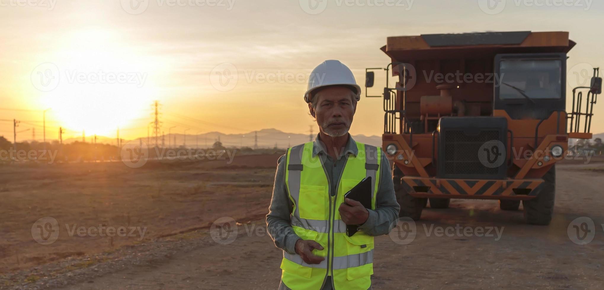 senior ingenieur die een veiligheidsvest en helm draagt, een digitale tablet vasthoudt in het bouwgebied, een oudere werknemer die aan het werk is, heeft een grote vrachtwagen als achtergrond foto