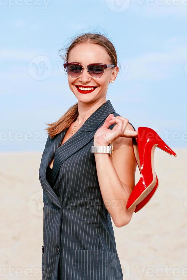portret van stijlvolle witte jonge vrouw in pak, zonnebril, staande met de glanzende hoge hakken in haar handen buiten foto