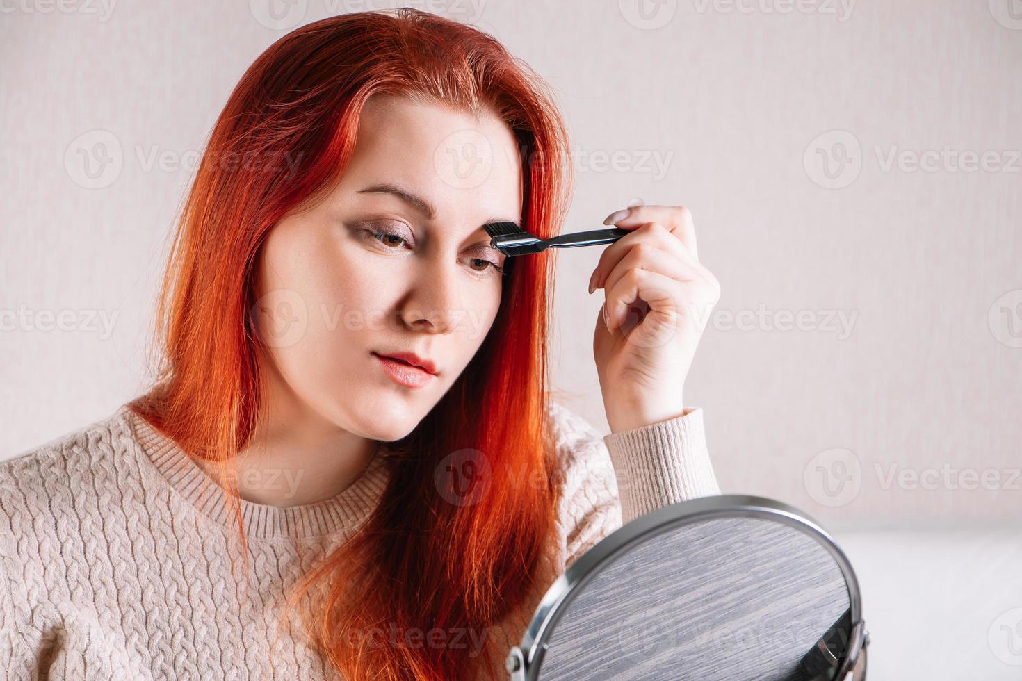 jonge vrouw met rood haar past make-up toe. dame kijkt in de spiegel en gebruikt cosmetica. foto