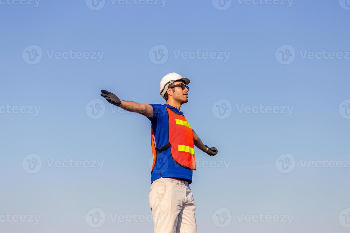 succesvolle jonge man met armen wijd open, gelukkige ingenieur voorman in veiligheidshelm en veiligheidsvest praat over blauwe hemelachtergrond foto