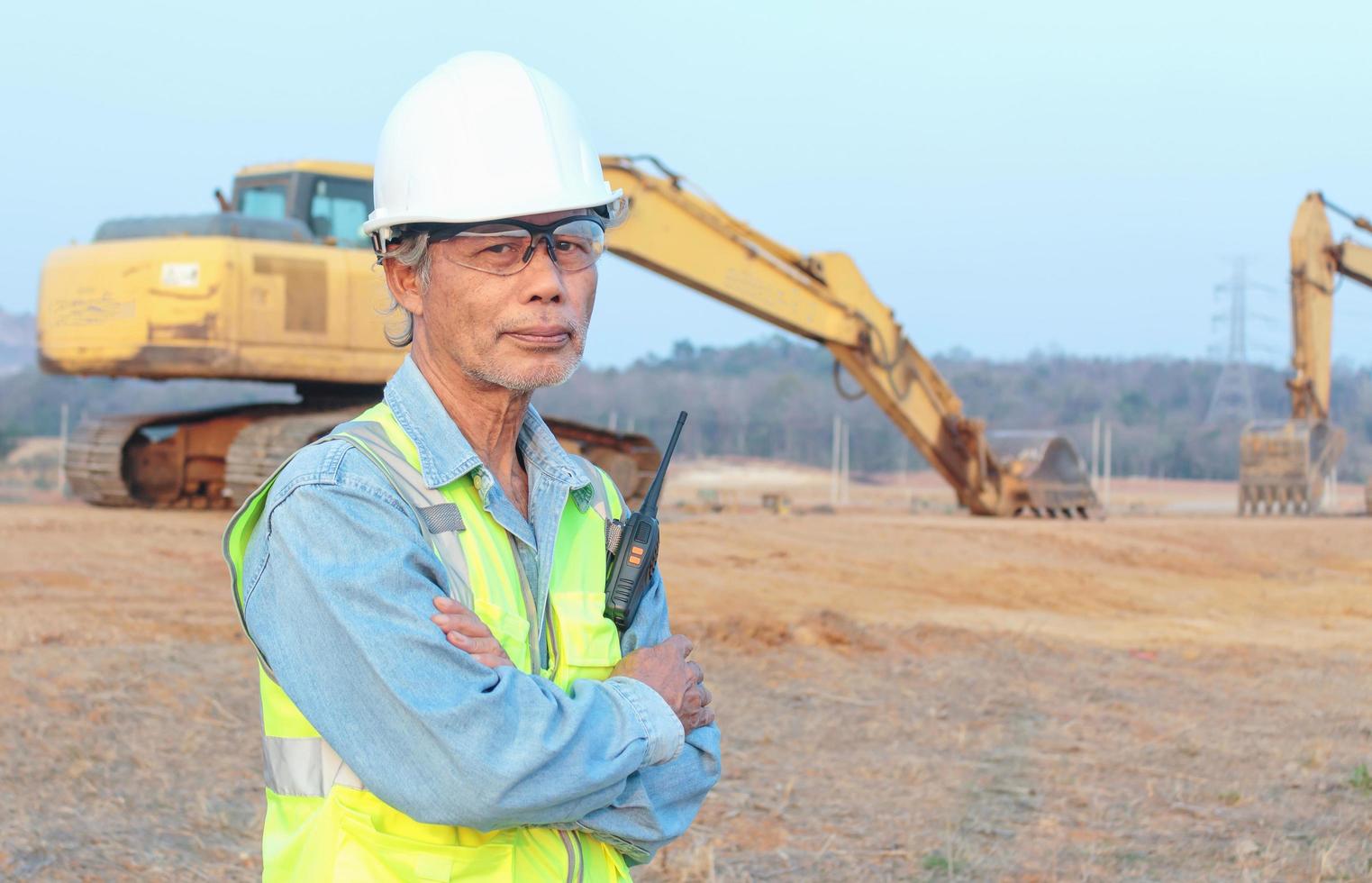 portret van een aziatische mannelijke senior ingenieur met een vest en helm inspecteert civiele werken, een voorman van middelbare leeftijd bestuurt een graafmachine. foto