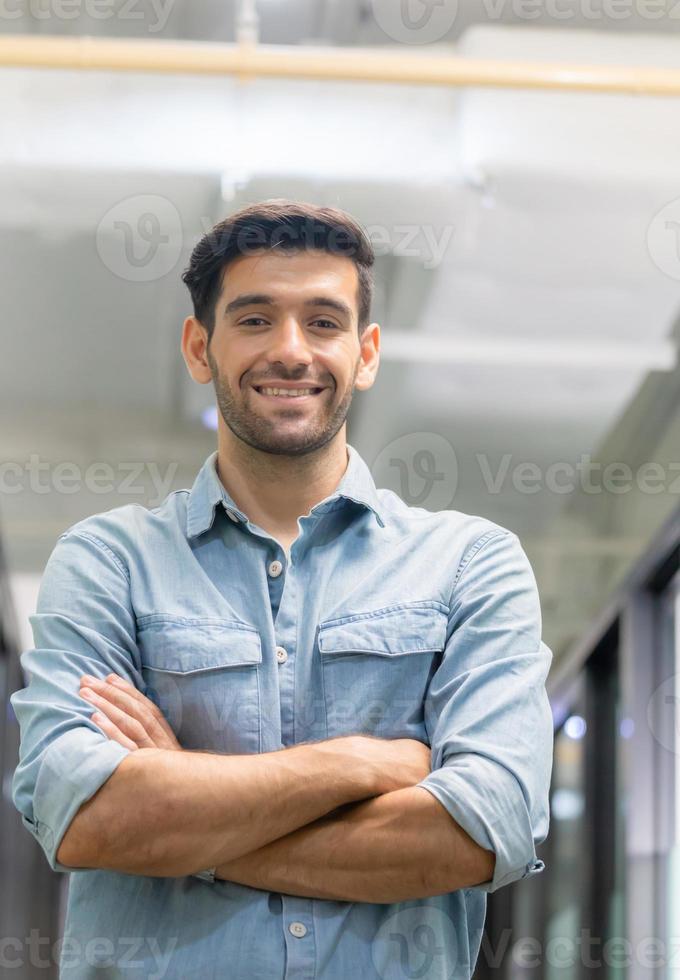 vrolijke casual jongeman die met gekruiste armen in kantoor staat foto