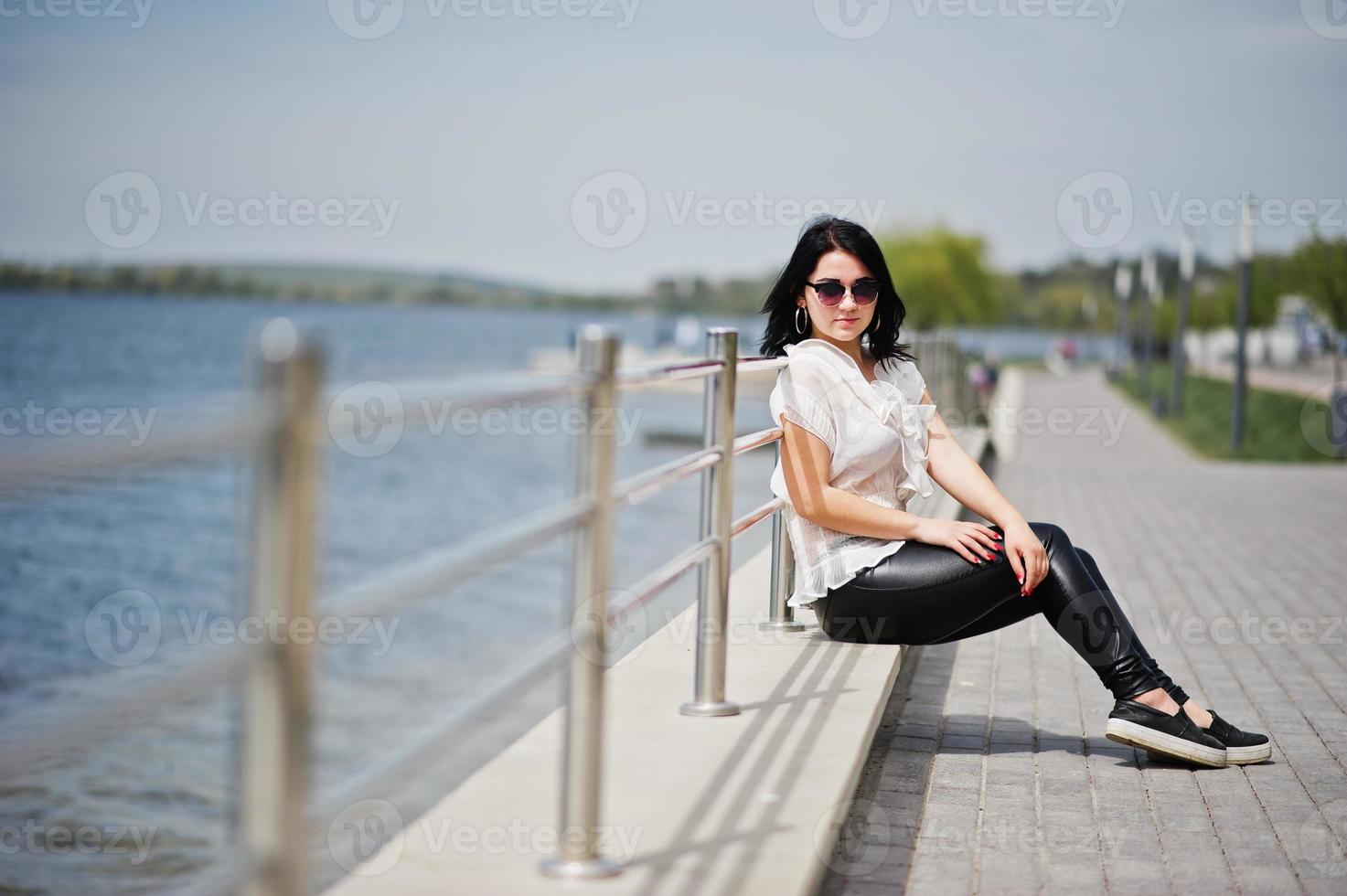 portret van brunette meisje op vrouwen leren broek en witte blouse, zonnebril, tegen ijzeren balustrades op het strand. foto