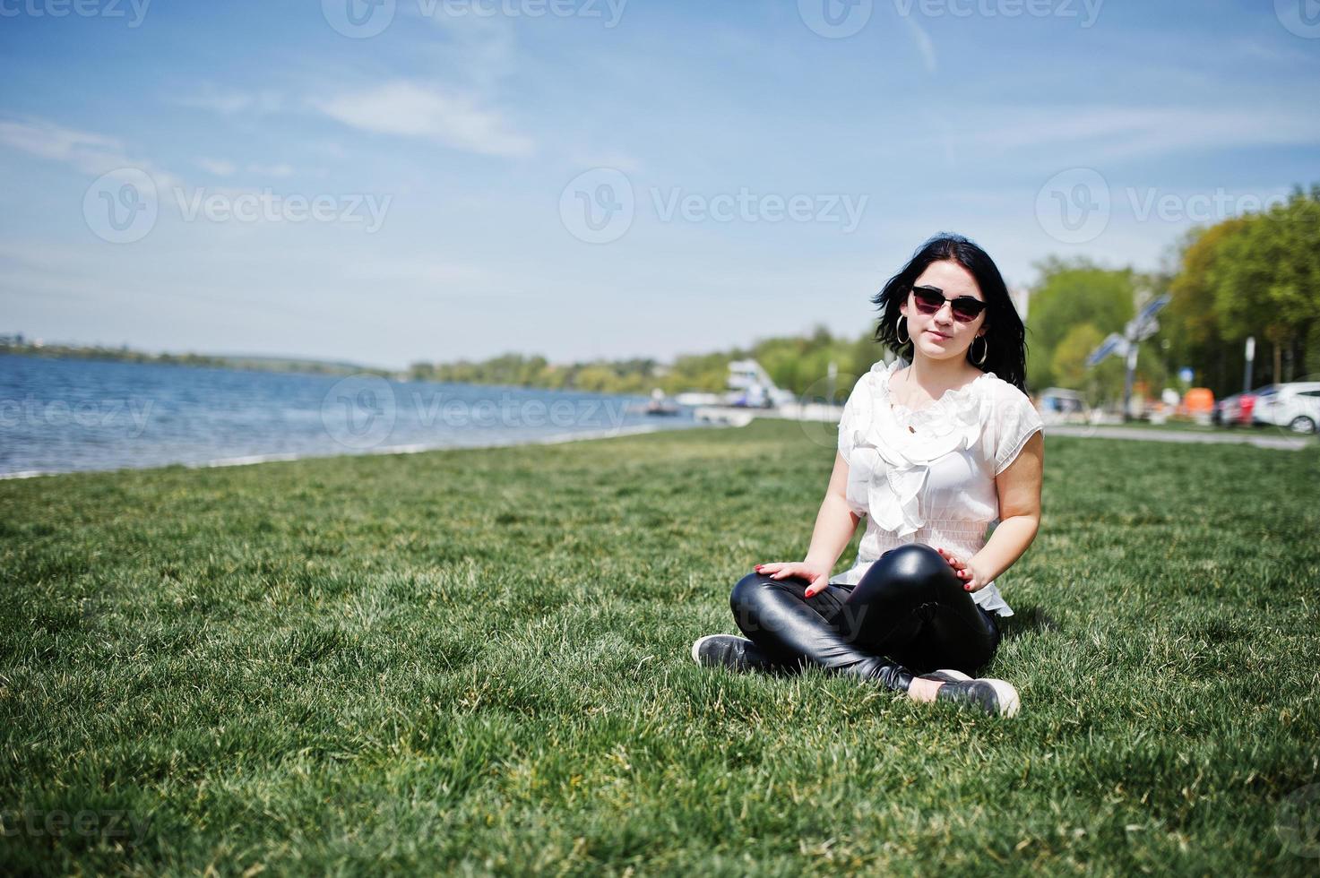 brunette meisje op leren damesbroek en witte blouse, zonnebril, zittend op geen gras tegen het strand van het meer. foto