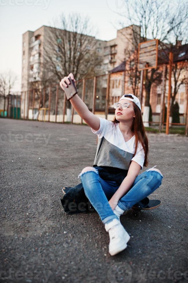 jonge stedelijke tienermeisje met skateboard, draag een bril, pet en gescheurde spijkerbroek op het sportveld van de werf bij zonsondergang selfie maken op de telefoon. foto