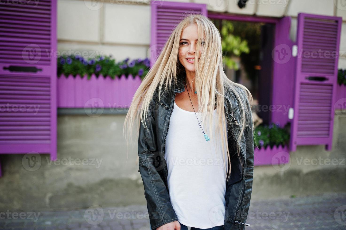 stijlvolle blonde vrouw draagt spijkerbroek en jas op straat tegen paars raam. mode stedelijk model portret. foto