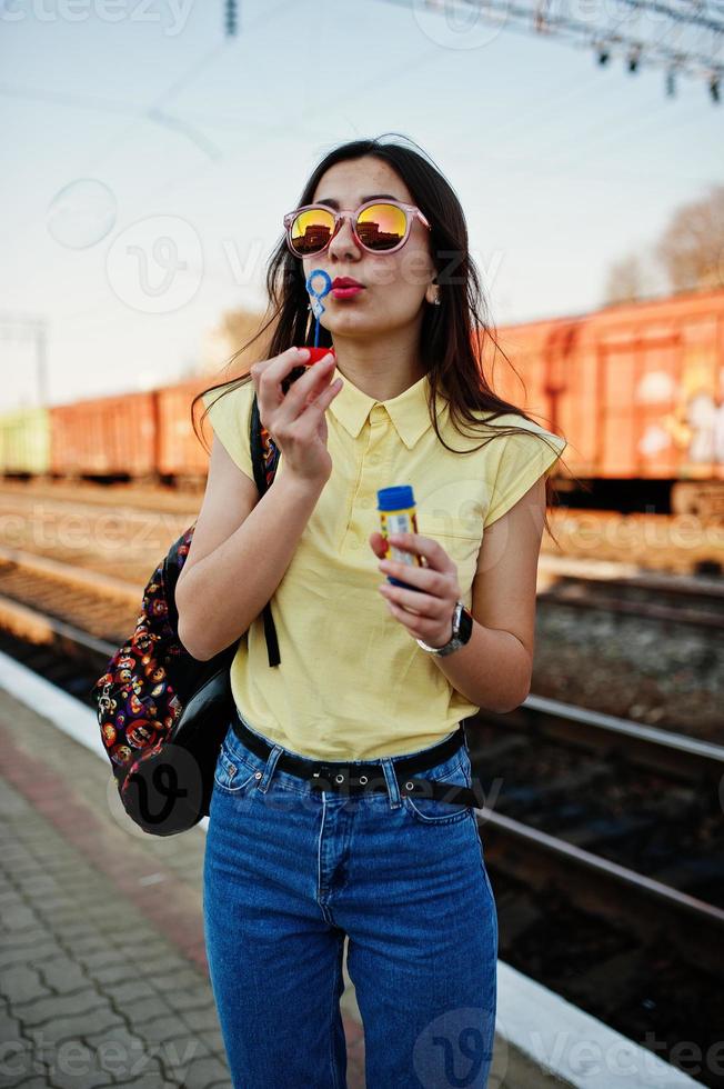 jong tienermeisje dat op het perron van het treinstation staat en zeepbellen blaast, draagt een geel t-shirt, jeans en zonnebril, met rugzak. foto