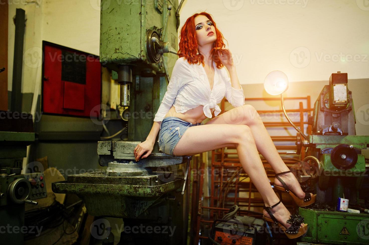 roodharige meisjesslijtage op korte denim shorts en witte blouse geposeerd op industriële machine in de fabriek. foto