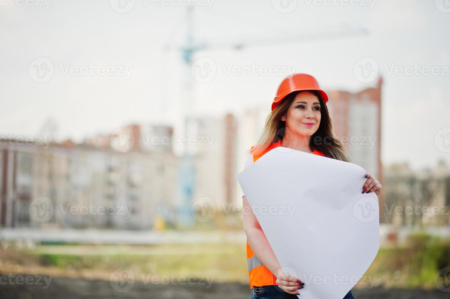 ingenieur bouwer vrouw in uniform vest en oranje beschermende helm houdt zakelijk papier tegen nieuwe gebouwen met kraan. foto