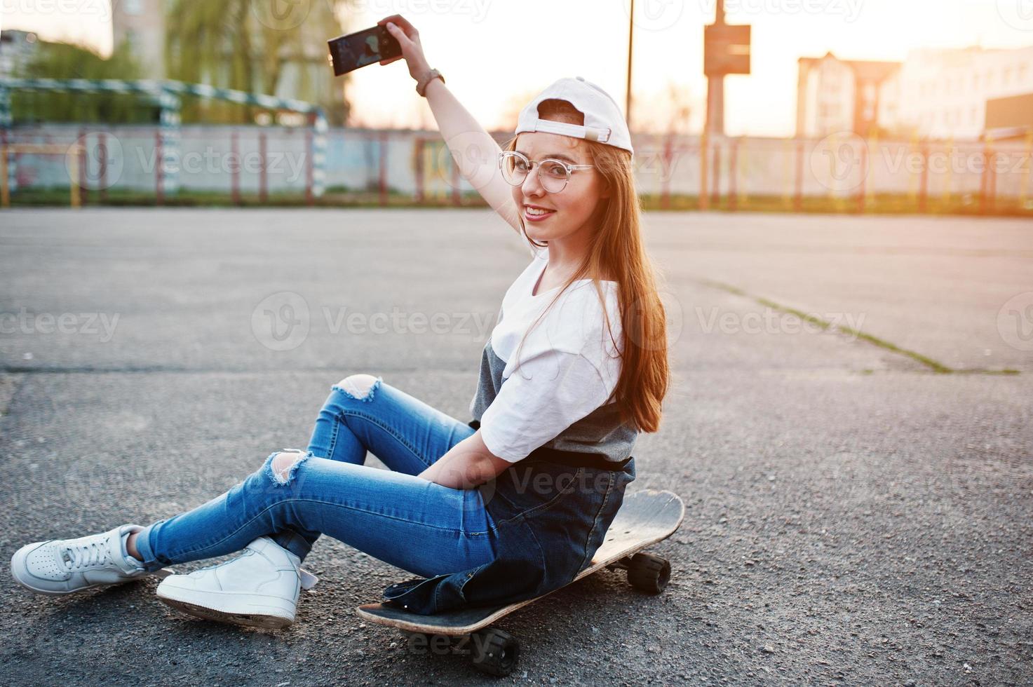 jonge stedelijke tienermeisje met skateboard, draag een bril, pet en gescheurde spijkerbroek op het sportveld van de werf bij zonsondergang selfie maken op de telefoon. foto