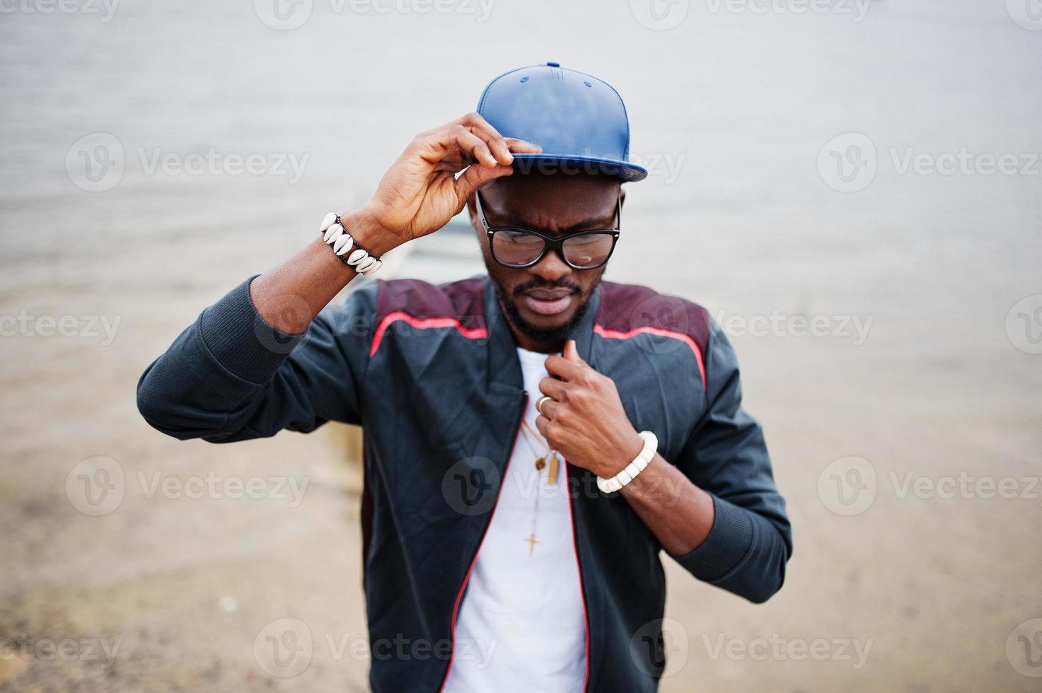 portret van stijlvolle Afro-Amerikaanse man op sportkleding, pet en bril tegen meer met boot. zwarte mannen model mode. foto