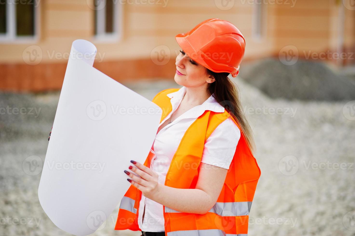 ingenieur bouwer vrouw in uniform vest en oranje beschermende helm houdt zakelijk papier tegen nieuwbouw. eigendom woonblok thema. foto