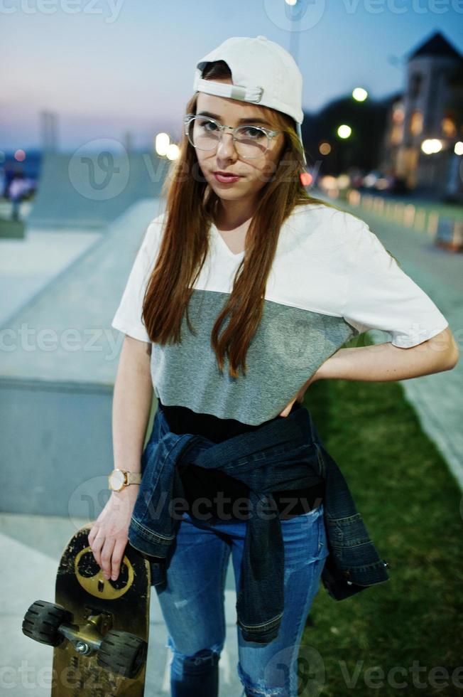 jonge stedelijke tienermeisje met skateboard, draag een bril, pet en gescheurde spijkerbroek in het skatepark op de avond. foto