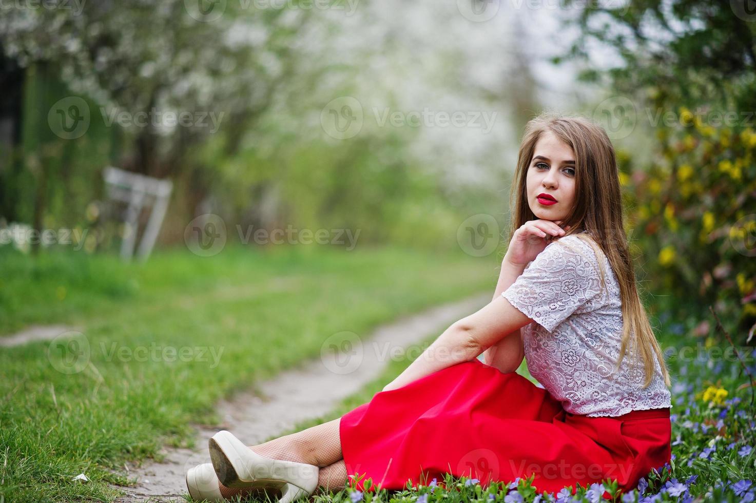 portret van sitiing mooi meisje met rode lippen op lente bloesem tuin op gras met bloemen, slijtage op rode jurk en witte blouse. foto