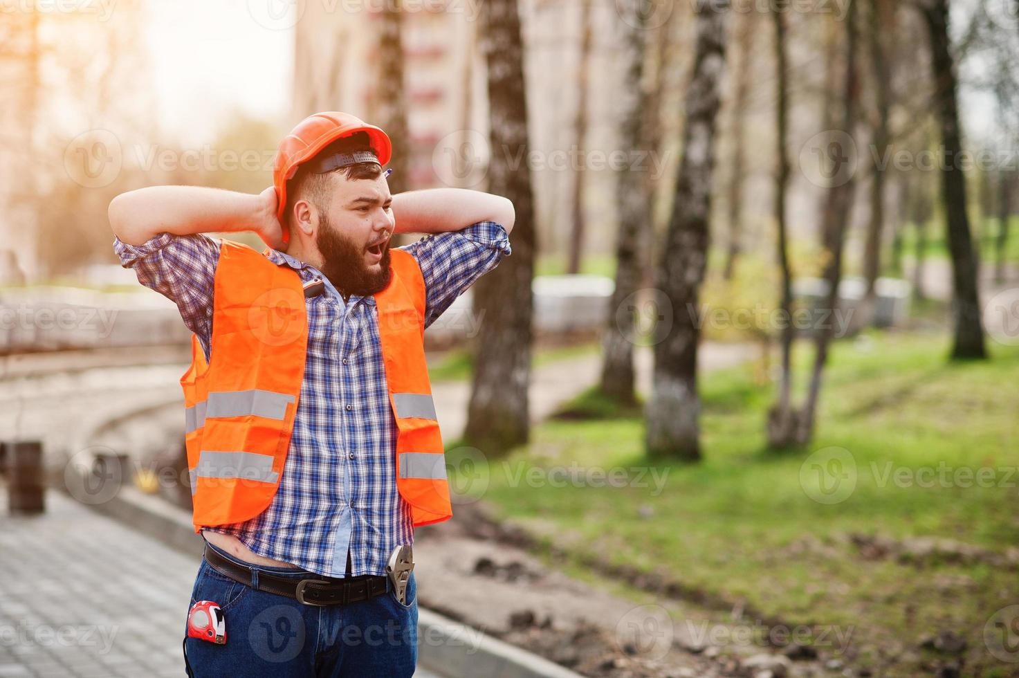 portret van luie gegaap brutale baard werknemer man pak bouwvakker in veiligheid oranje helm tegen bestrating. foto