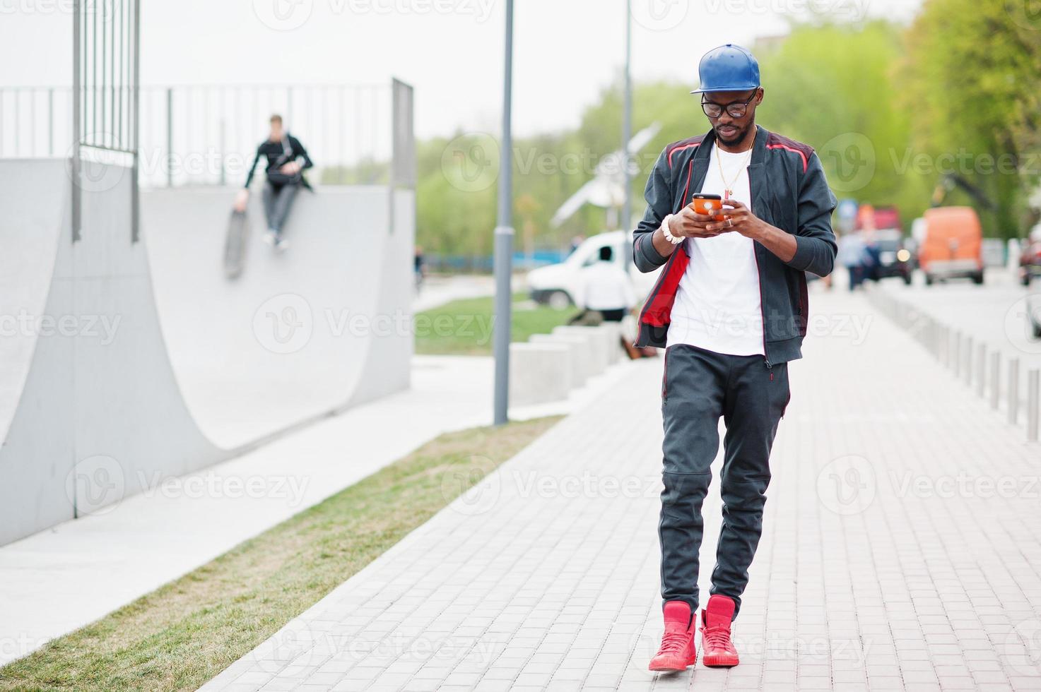portret van een stijlvolle Afro-Amerikaanse man op sportkleding, pet en bril die op zijn telefoon kijkt. zwarte mannen model lopen in skatepark. foto