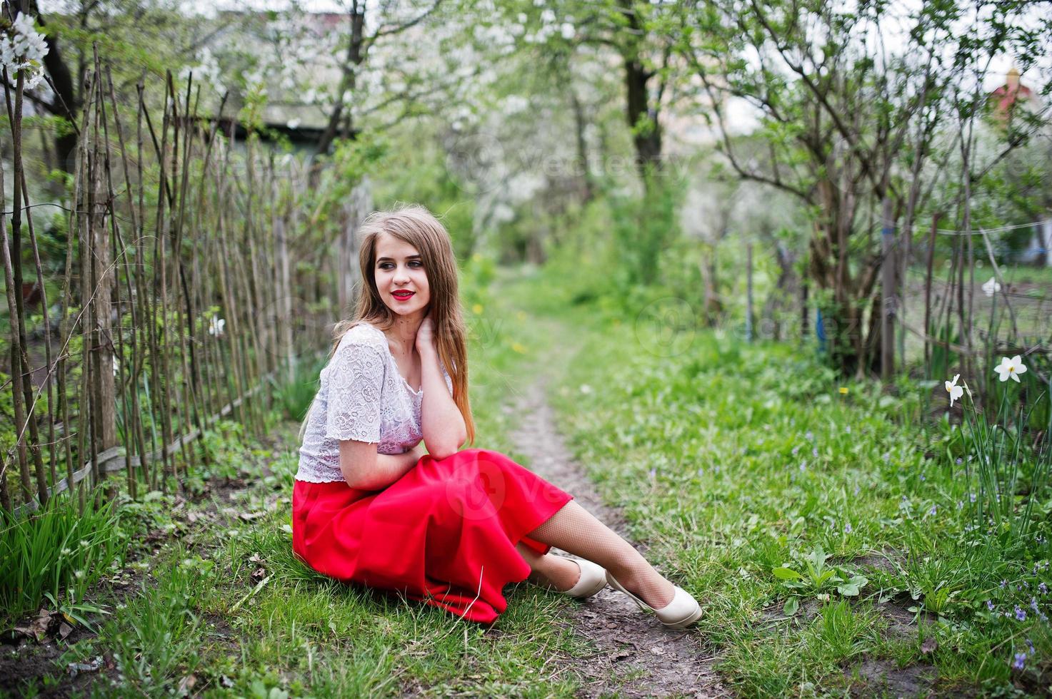 portret van een mooi meisje met rode lippen in de lentebloesemtuin op groen gras, een rode jurk en een witte blouse dragen. foto