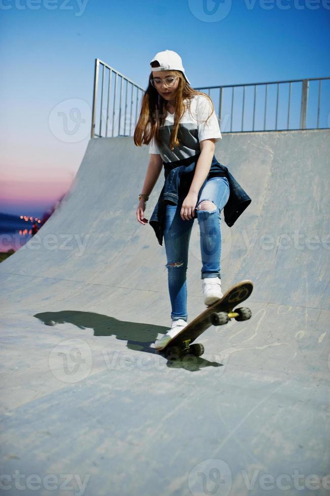 jonge stedelijke tienermeisje met skateboard, draag een bril, pet en gescheurde spijkerbroek in het skatepark op de avond. foto