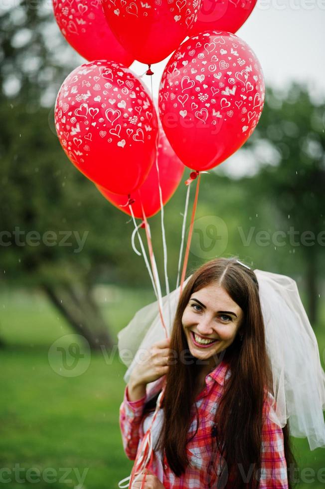 portret van brunette meisje op geruit hemd, jeans en sluier met veel rode ballonnen op vrijgezellenfeest. foto