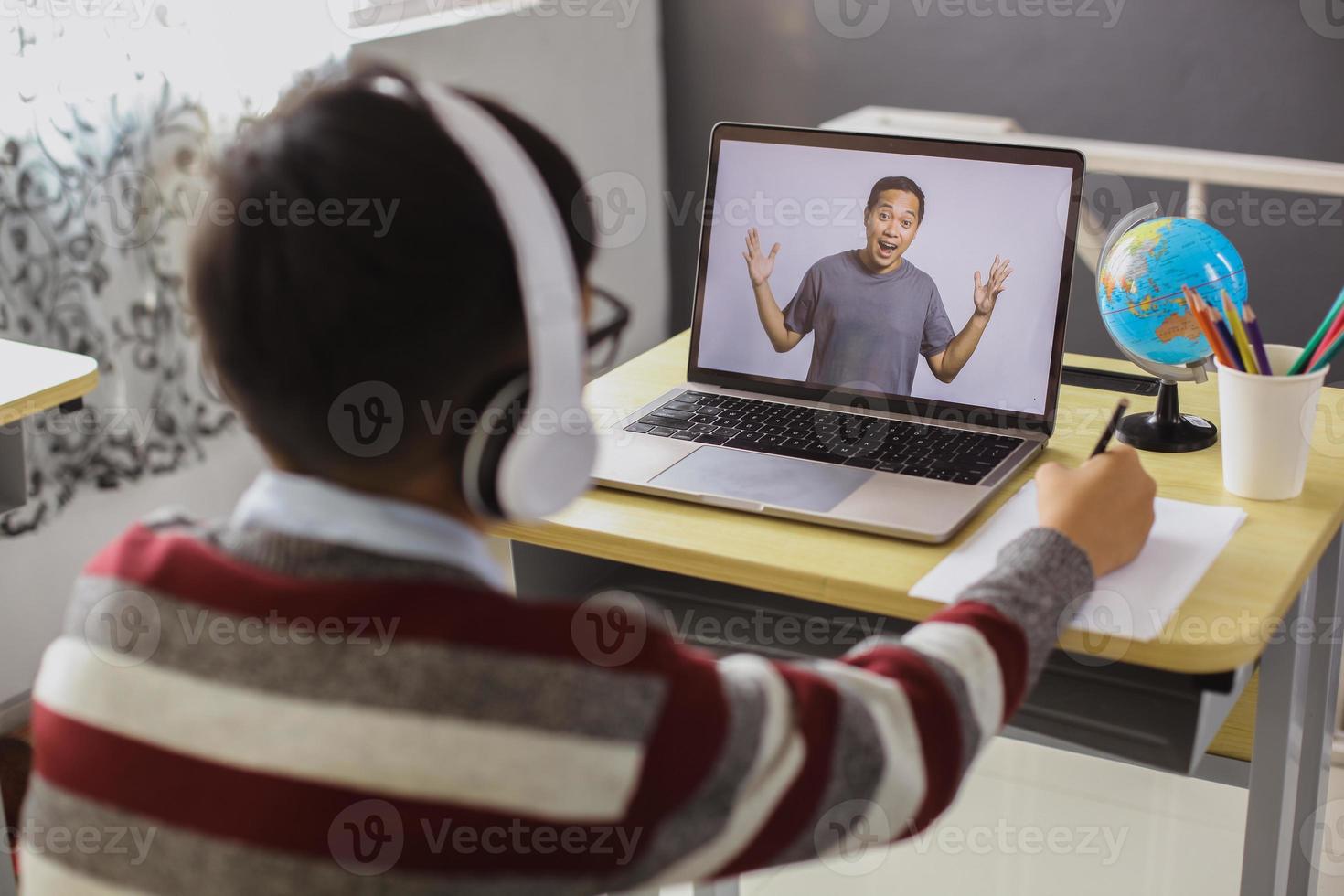 een schooljongen studeert en kijkt naar zijn leraar op een laptop terwijl hij op papier schrijft foto