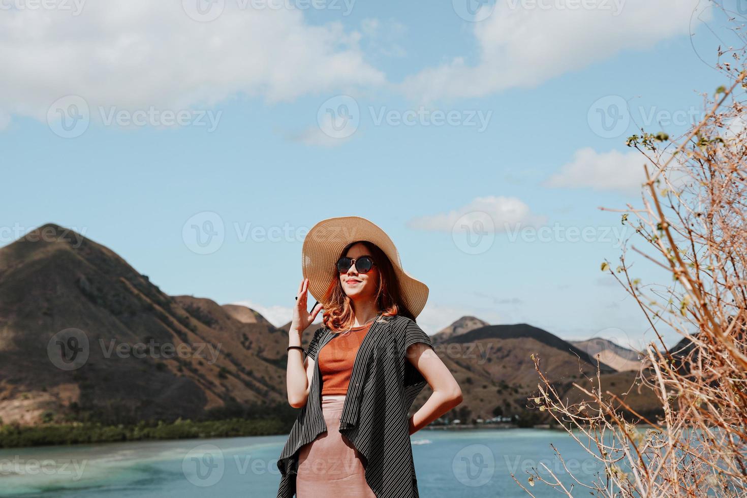 glimlachende aziatische vrouwen in zomerhoed en zonnebril terwijl ze poseren met een achtergrond van zeegezicht en heuvels bij labuan bajo foto