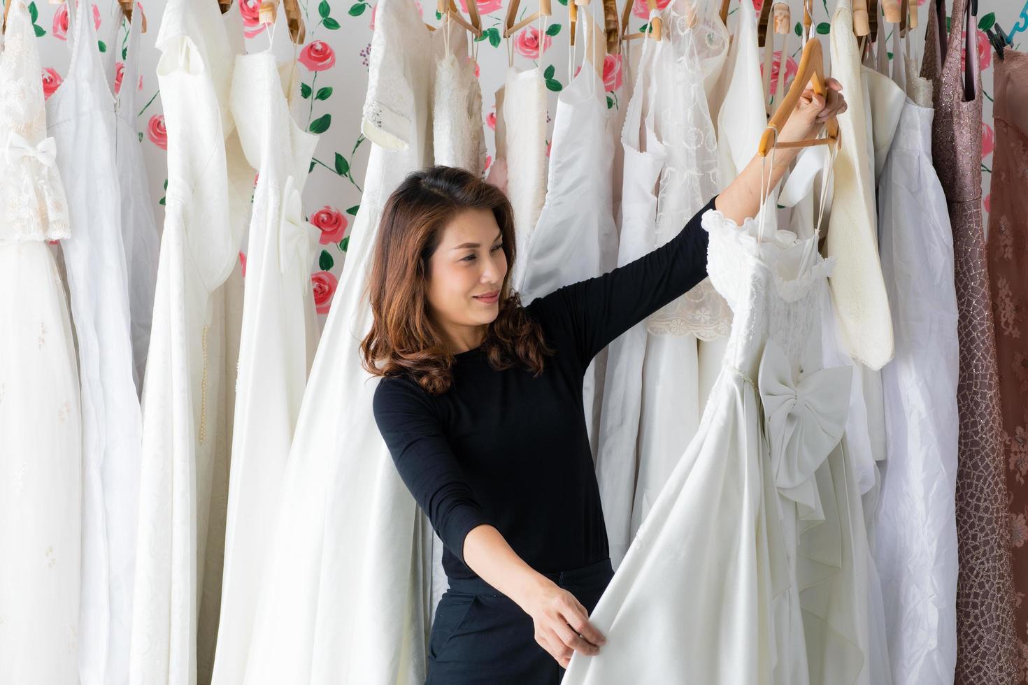 glamour aziatische vrouw bruid bruiloft winkeleigenaar werkt in haar studio, selecteert en beoordeelt kleding en bruidsjurk met een glimlach en geluk. ontwerper en zakelijk ondernemer concept foto